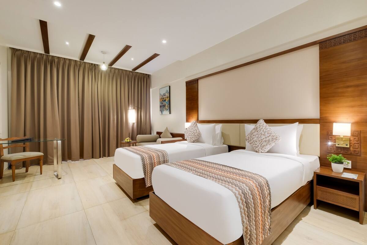 A hotel room with two beds, a desk, a chair, a bedside table with a lamp, and a wall-mounted artwork.
