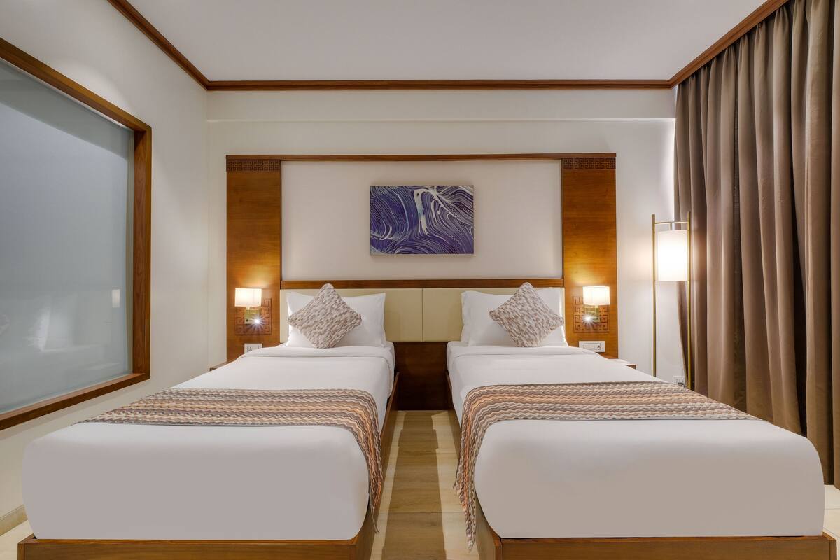 A hotel room with two beds, a large window, a wall-mounted artwork, and a nightstand with a lamp.