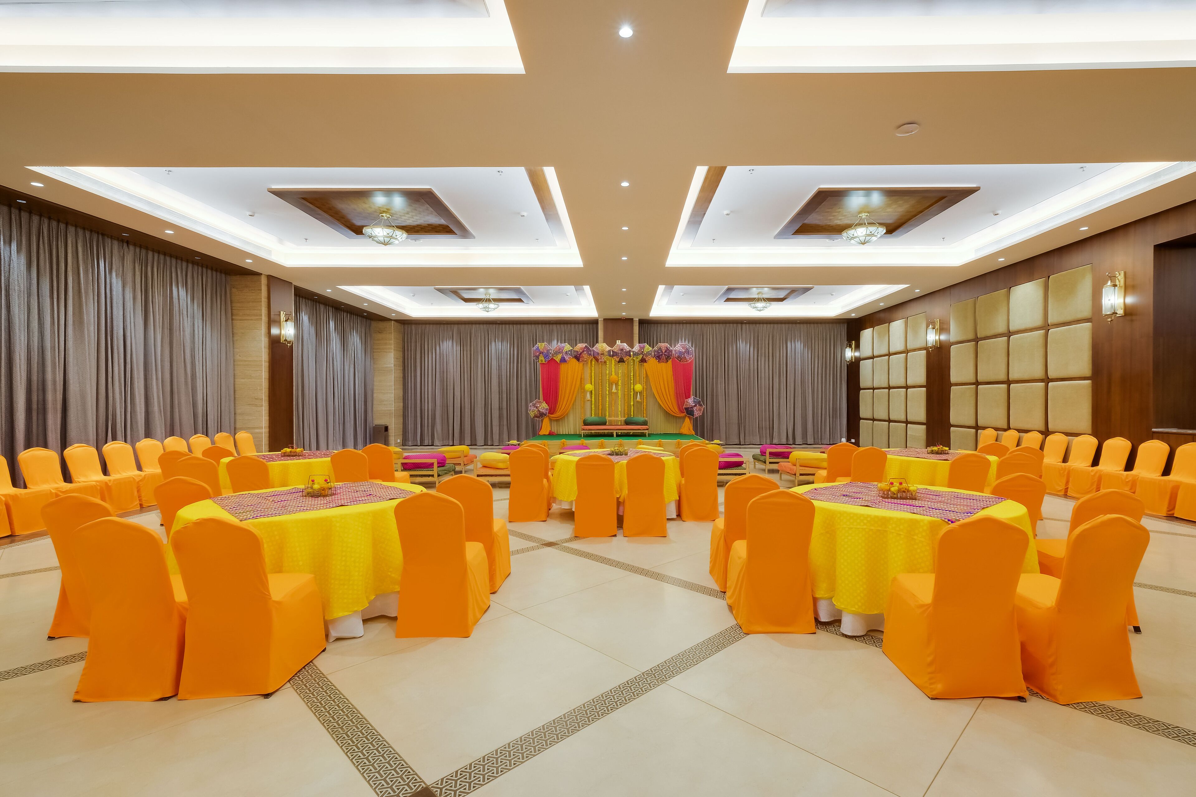 Un salón de banquetes con mesas redondas y sillas anaranjadas, un escenario con fondo floral e iluminación empotrada en el techo.