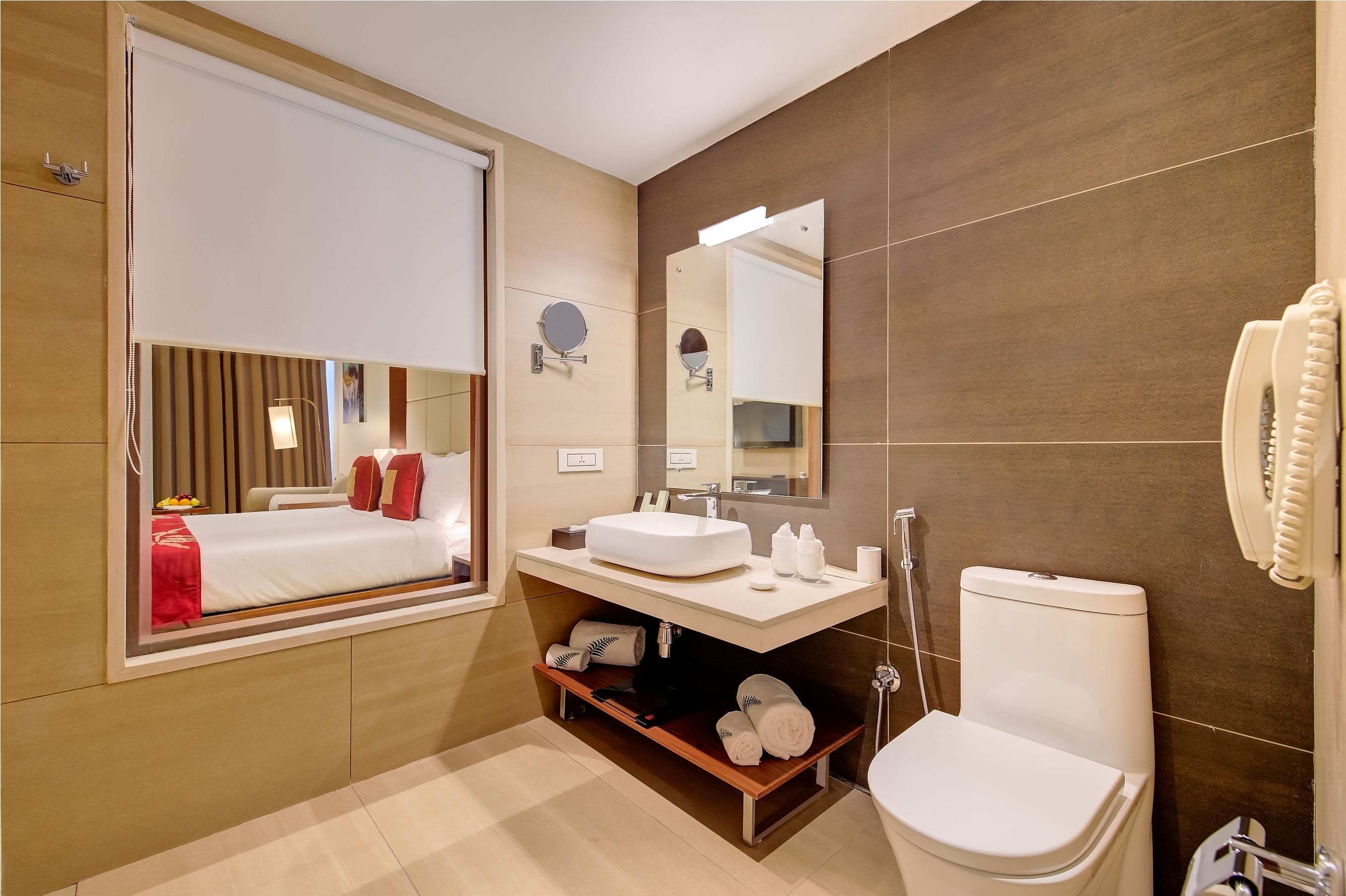 A modern bathroom with a large mirror, a sink, and a toilet. There is a window with blinds, a towel rack, and a shelf with towels.