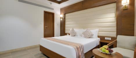 Habitación de hotel con una cama grande, un cabecero de madera, una pequeña mesa con fruta y una silla.