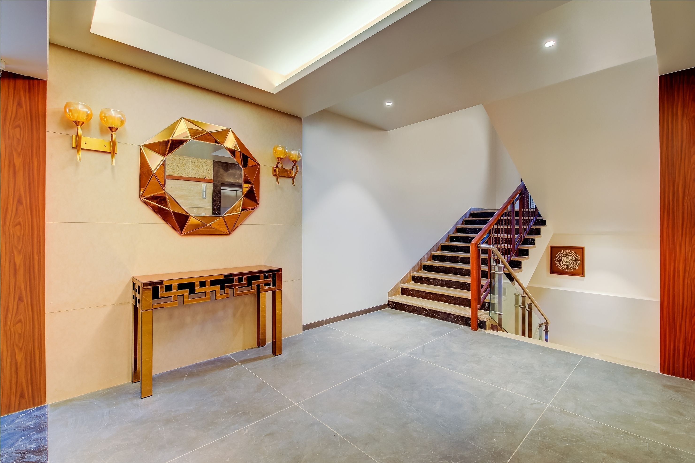 A modern interior with a geometric mirror, wall sconces, a console table, and a staircase.