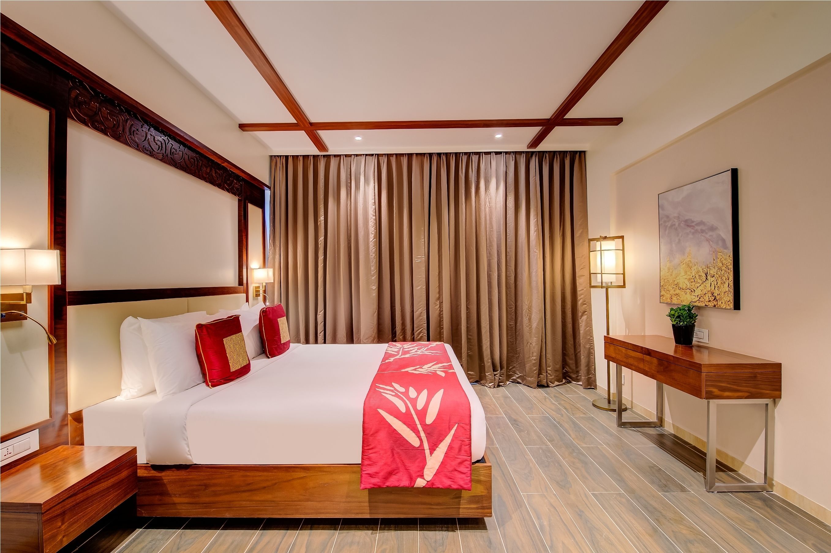 Une chambre d’hôtel avec un grand lit, une tête de lit en bois, une table de chevet, une commode, un bureau avec une lampe et une grande fenêtre avec des rideaux.