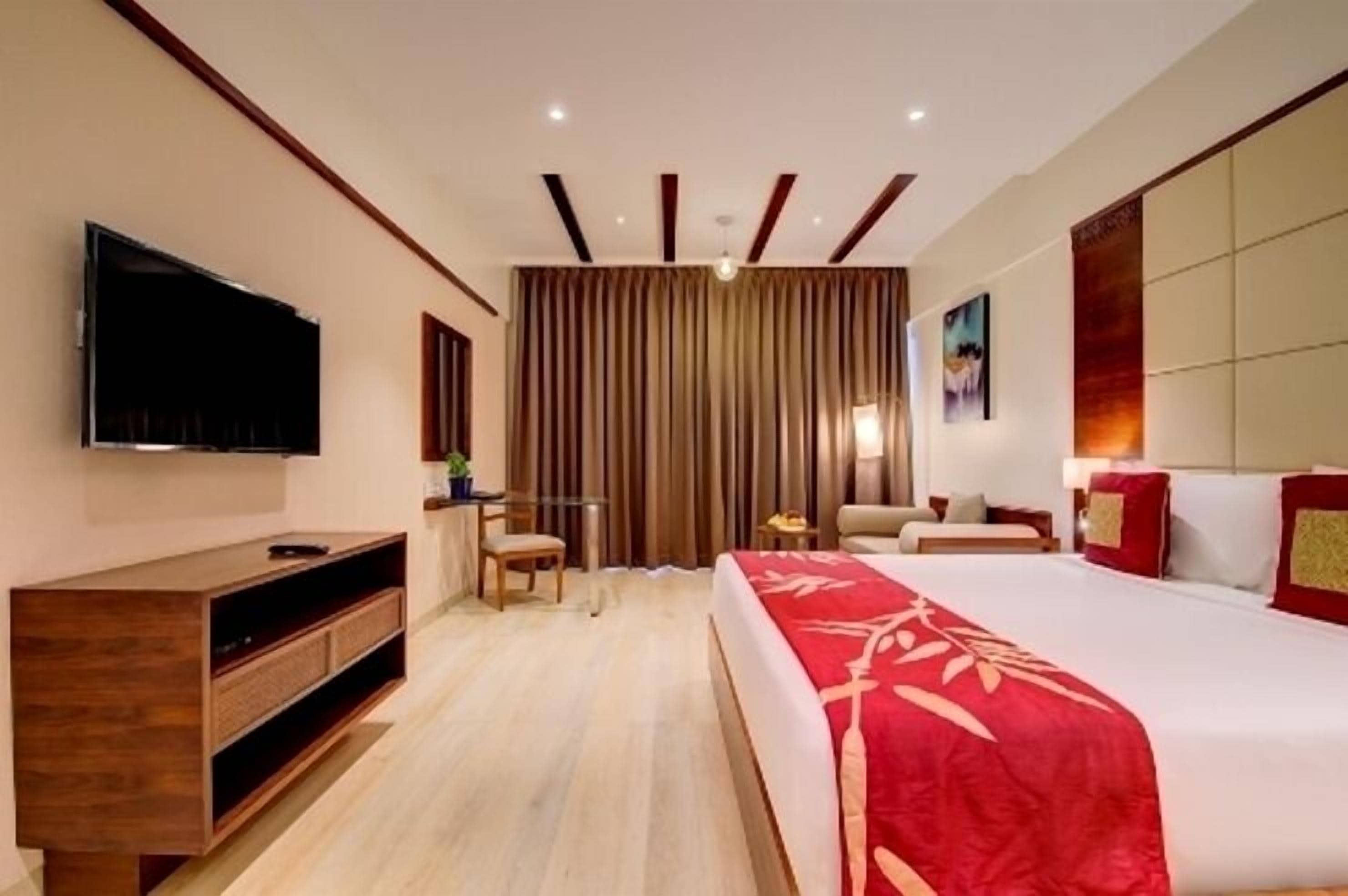 A hotel room with a large bed, a flat-screen TV mounted on the wall, a wooden cabinet, a desk with a chair, and a large window with curtains.