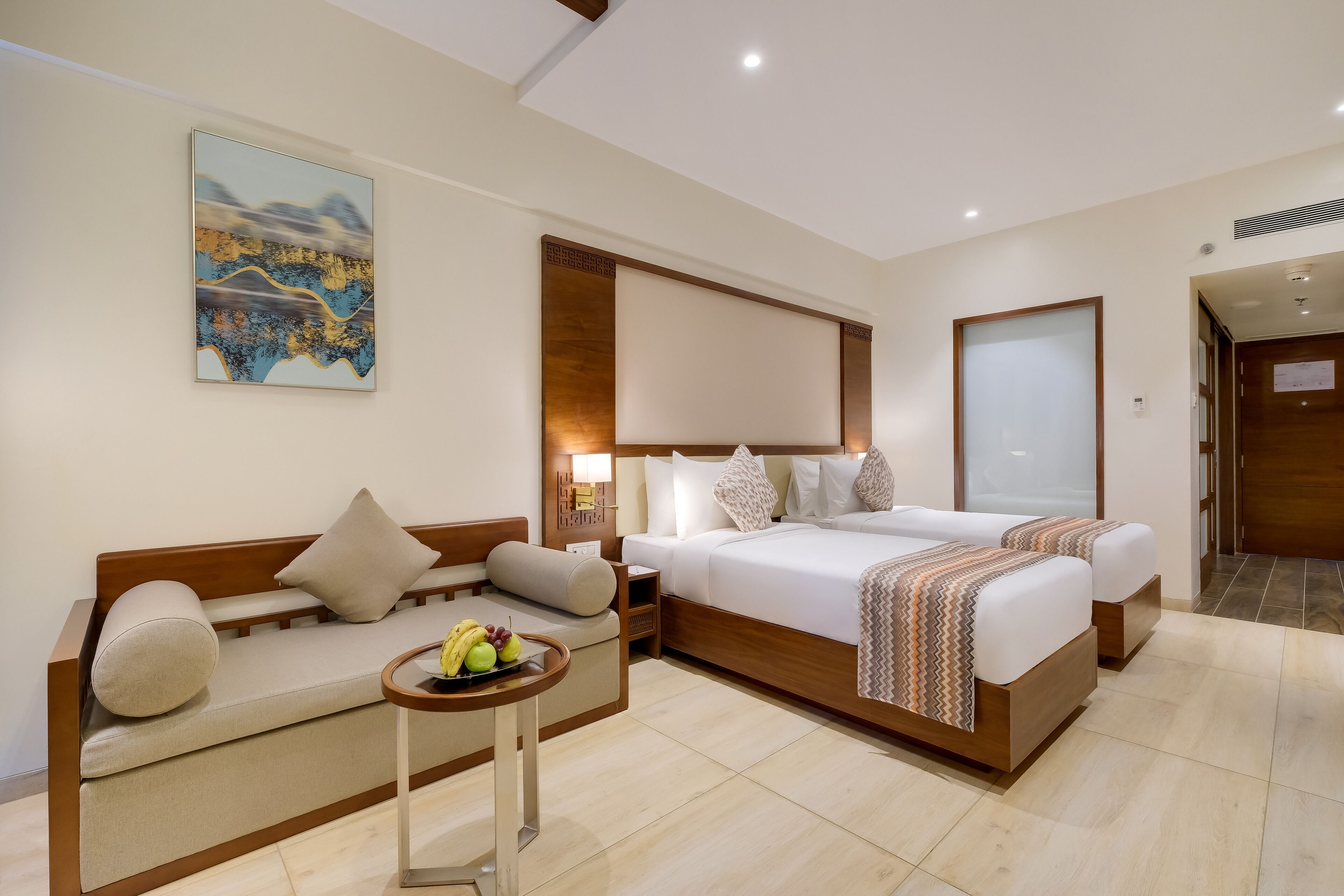 A hotel room with a bed, a sofa, a small table with fruit, and a painting on the wall.