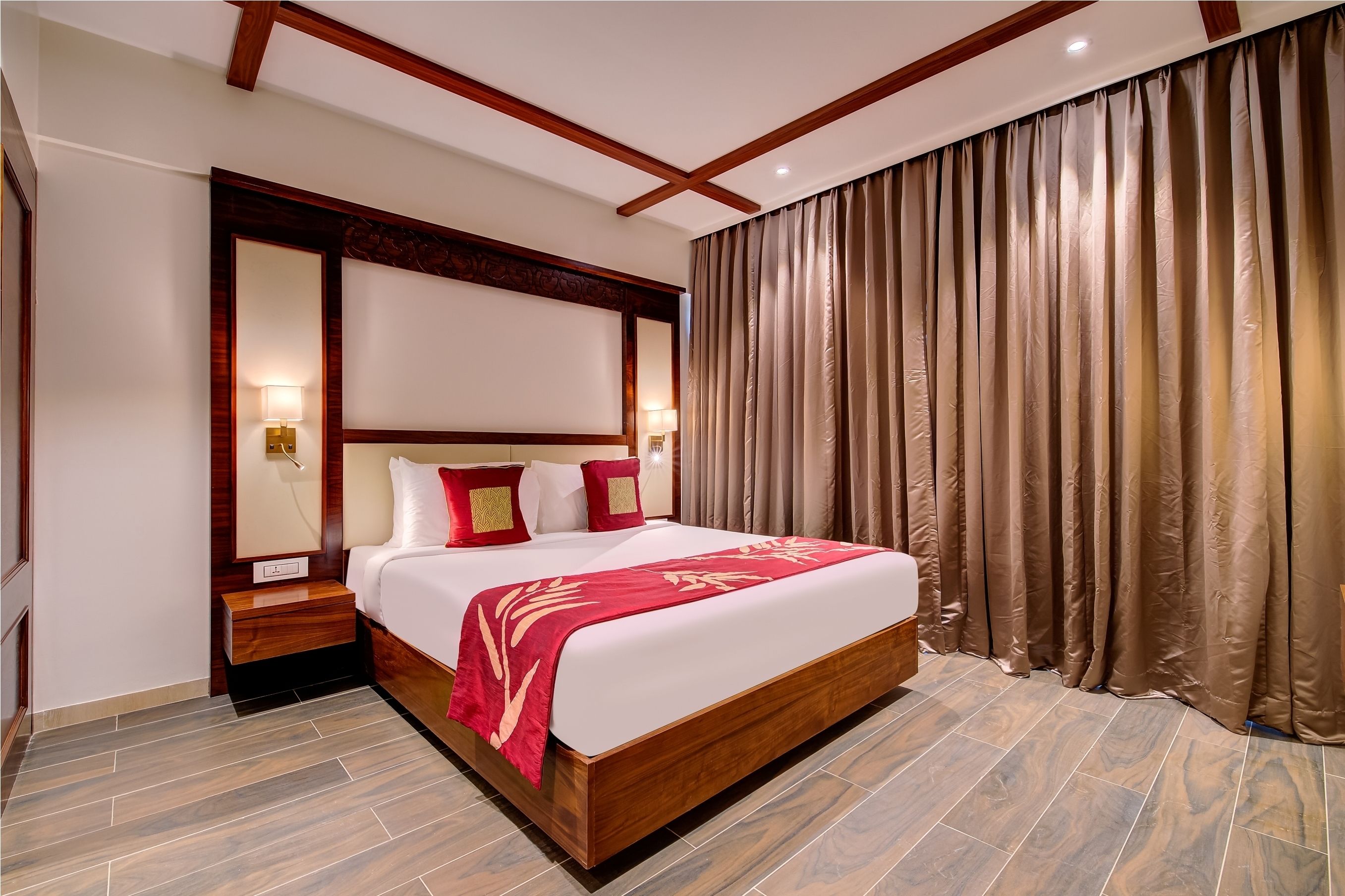 A hotel room with a wooden headboard, a large bed with red bedding, a nightstand, and a wall-mounted lamp.