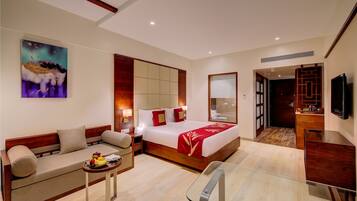 A modern hotel room with a bed, sofa, bedside table with fruit, a glass table, and a TV.