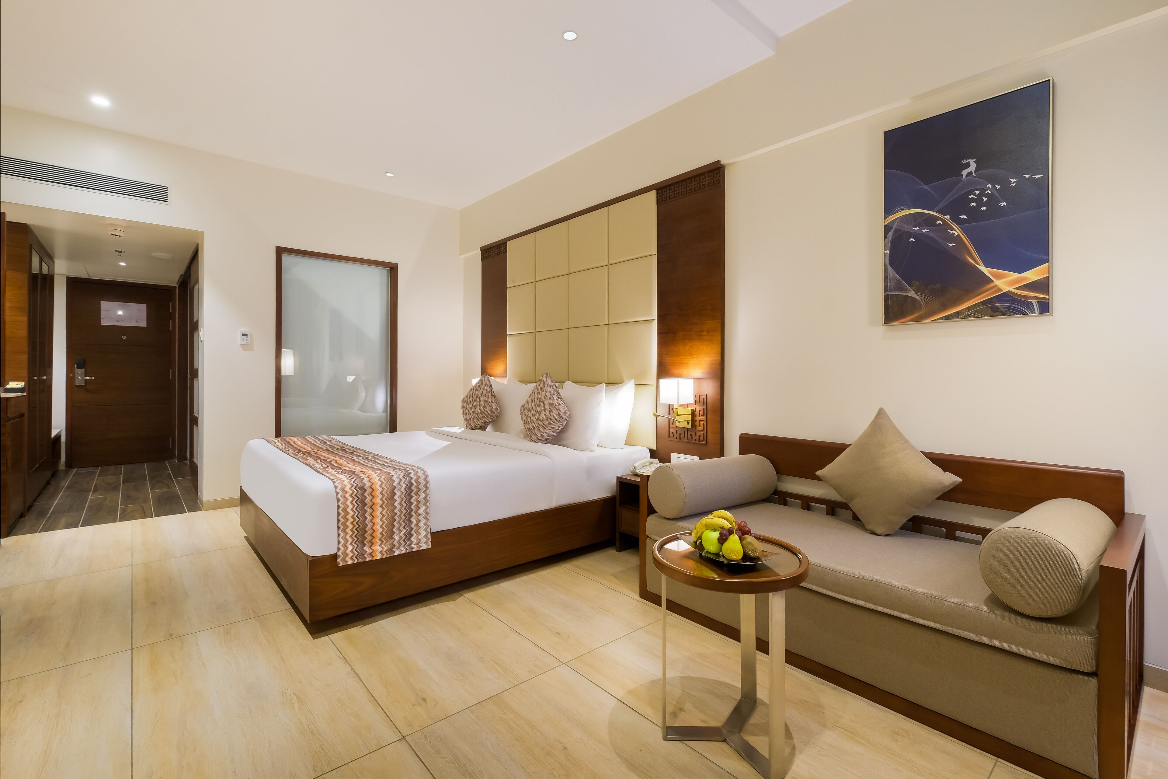 A hotel room with a large bed, a sofa, a small table with fruit, and a painting on the wall.