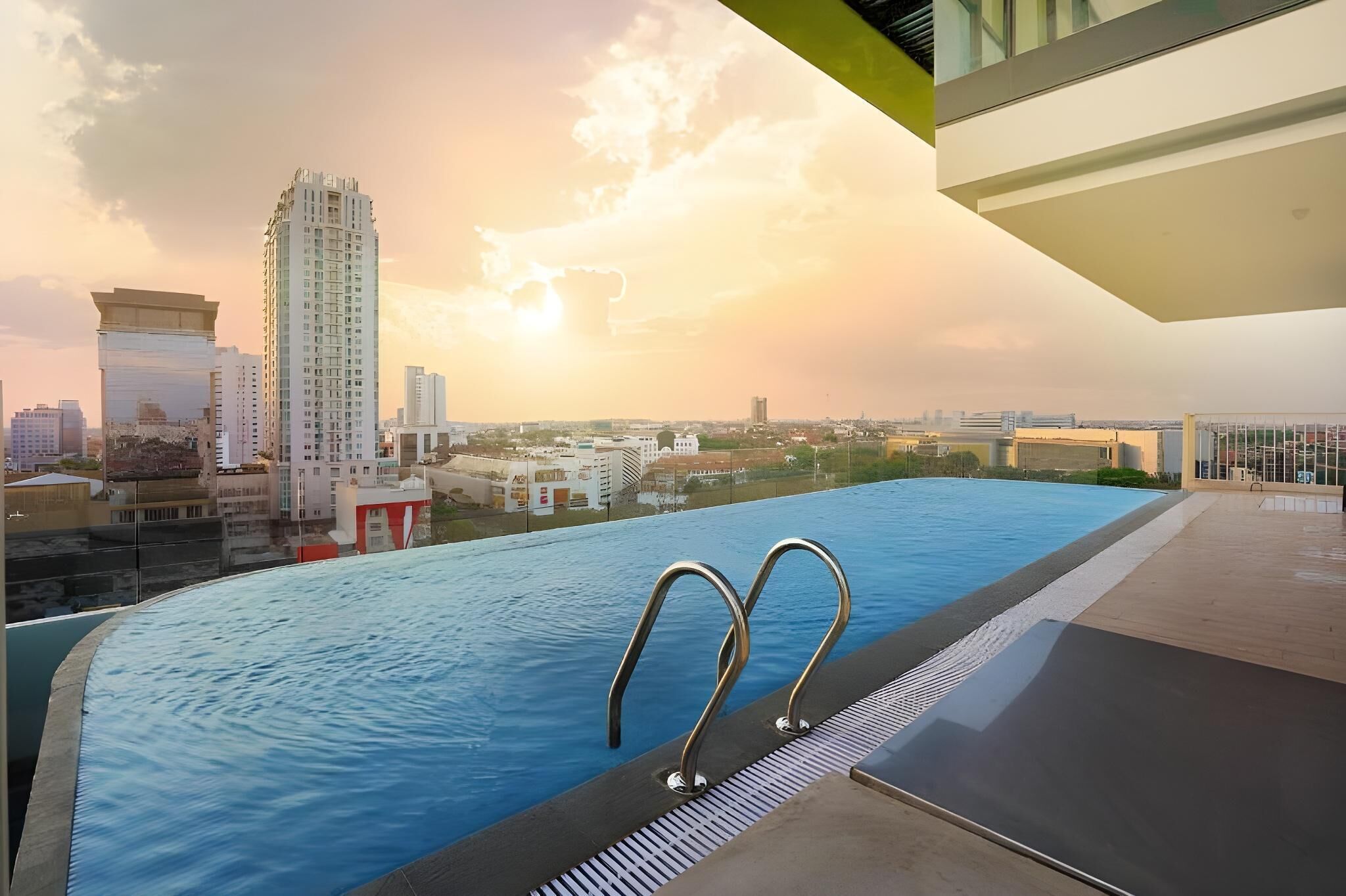 An infinity pool with metal railings, overlooking a cityscape with high-rise buildings during sunset.