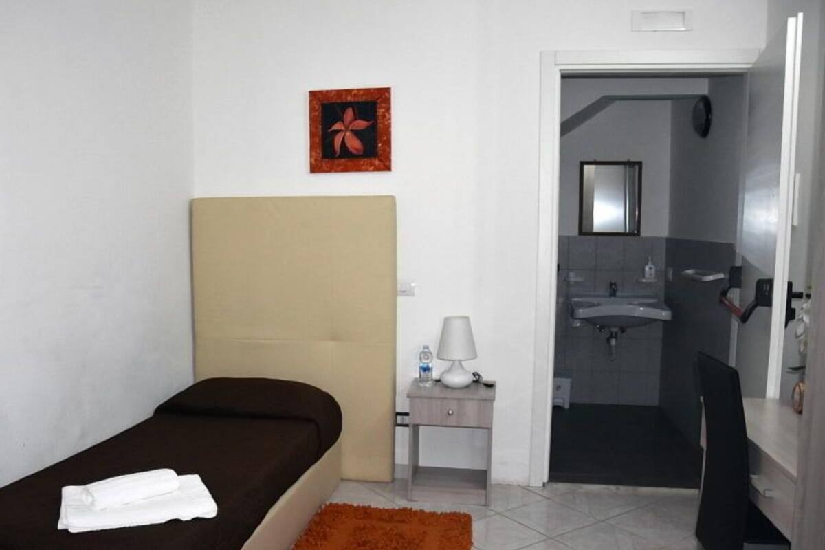 A small, clean room with a bed, a nightstand, a lamp, and a bathroom visible through an open door.