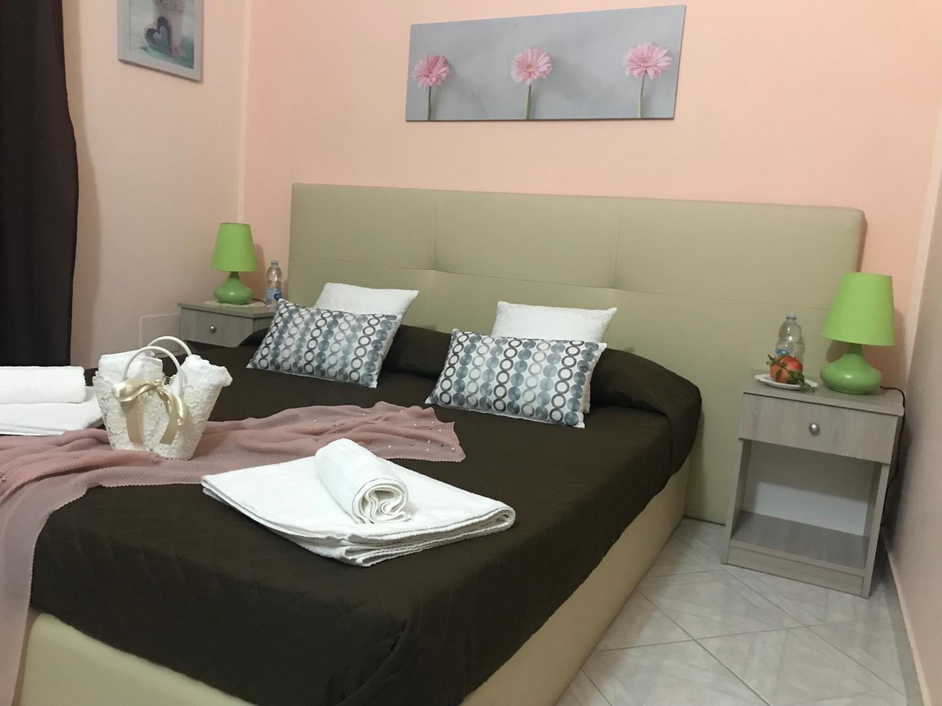 A neatly made bed with pillows, a folded towel, and a handbag on the side. There are two nightstands with lamps and a small table with a fruit and a bottle.