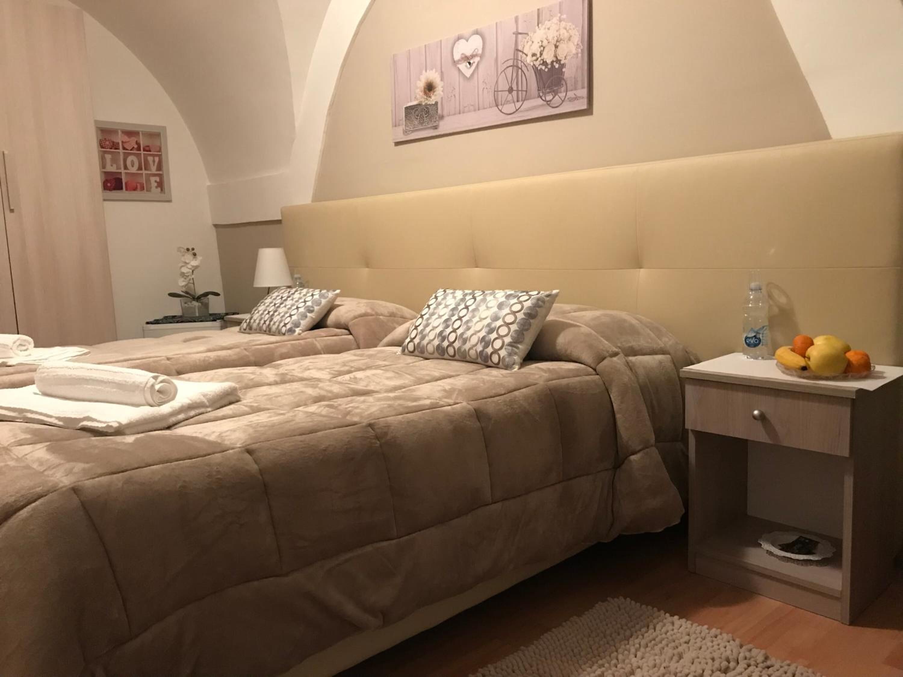 A hotel room with two beds, a nightstand with a water bottle and fruit, and a wall with a 'LOVE' sign and a bicycle painting.
