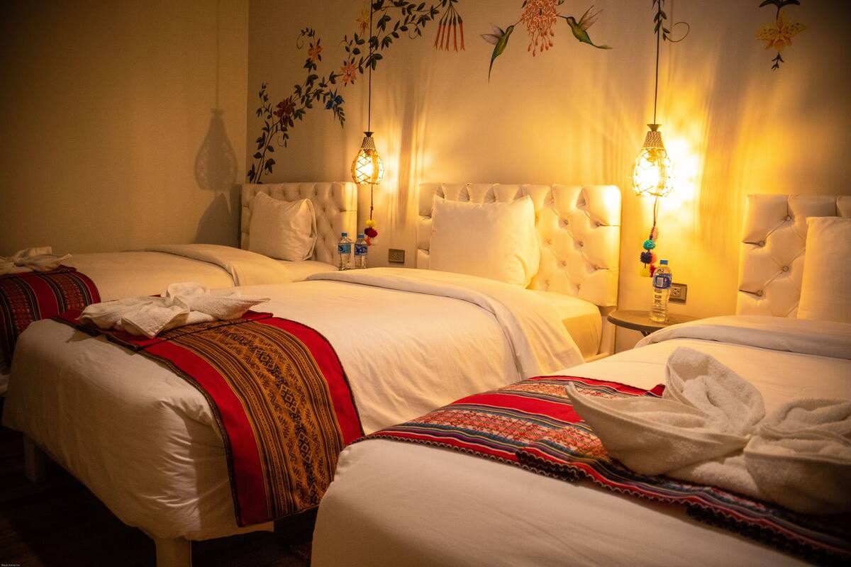 Une chambre d’hôtel avec deux lits, chacun recouvert d’une couette blanche et de couvertures rouges et dorées. Deux lampes murales décoratives sont fixées au mur.