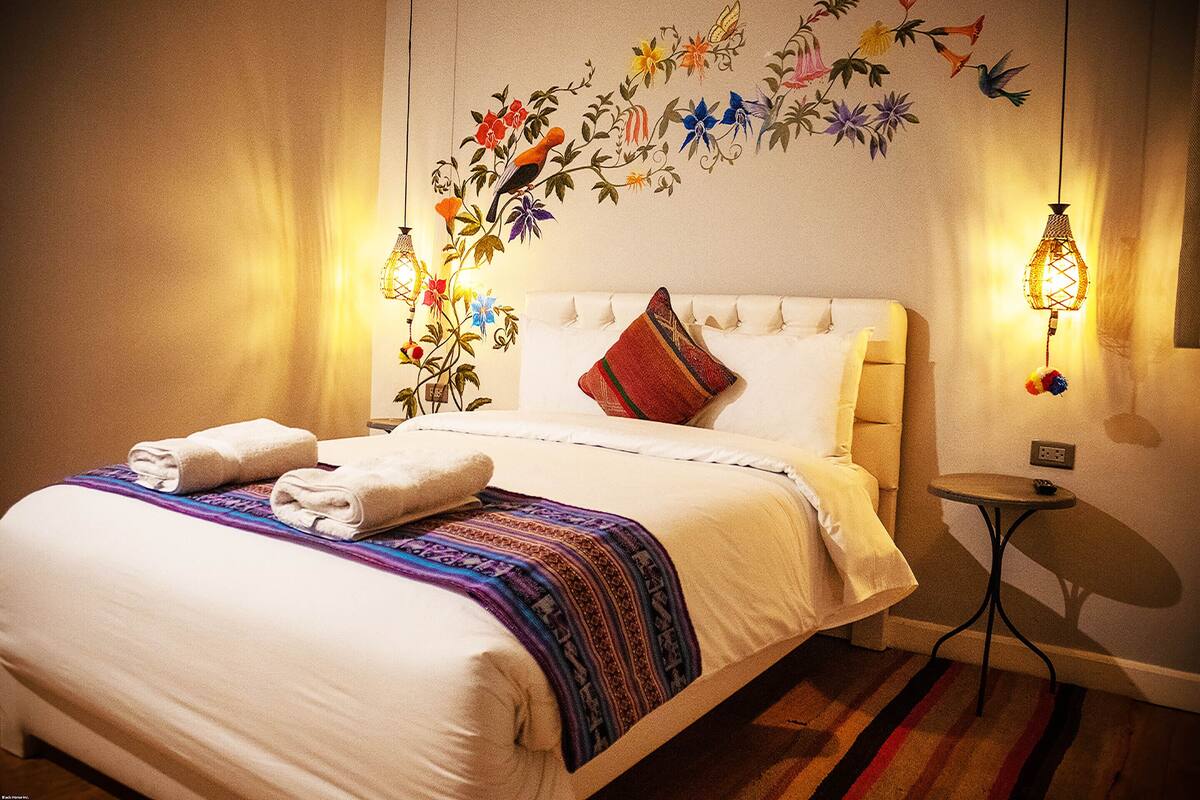 Un lit bien fait, agrémenté d’une couverture colorée et de deux serviettes, une fresque murale représentant des oiseaux et des fleurs, et deux lampes fixées au mur.