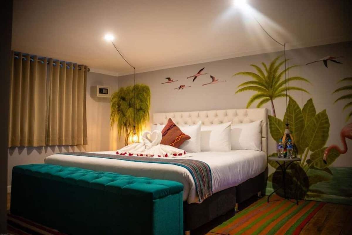 Une chambre avec un grand lit, un pouf vert, un climatiseur fixé au mur et une fresque murale sur le thème des tropiques.