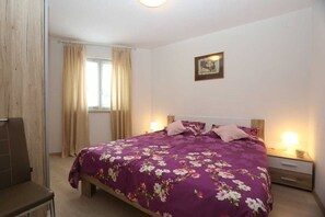 Une chambre à coucher comprenant un lit, deux tables de chevet, une chaise, une fenêtre avec des rideaux et un tableau accroché au mur.