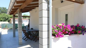 Zona exterior coberta amb una taula i caires, una paret blanca amb una finestra, i una jardineria amb flors roses.