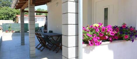 A covered outdoor area with a table and chairs, a white wall with a window, and a flower box with pink flowers.