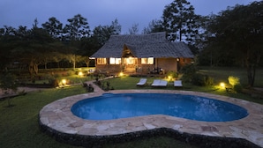 Uma casa com piscina cercada por ladrilhos de pedra, gramado e algumas cadeiras de descanso.