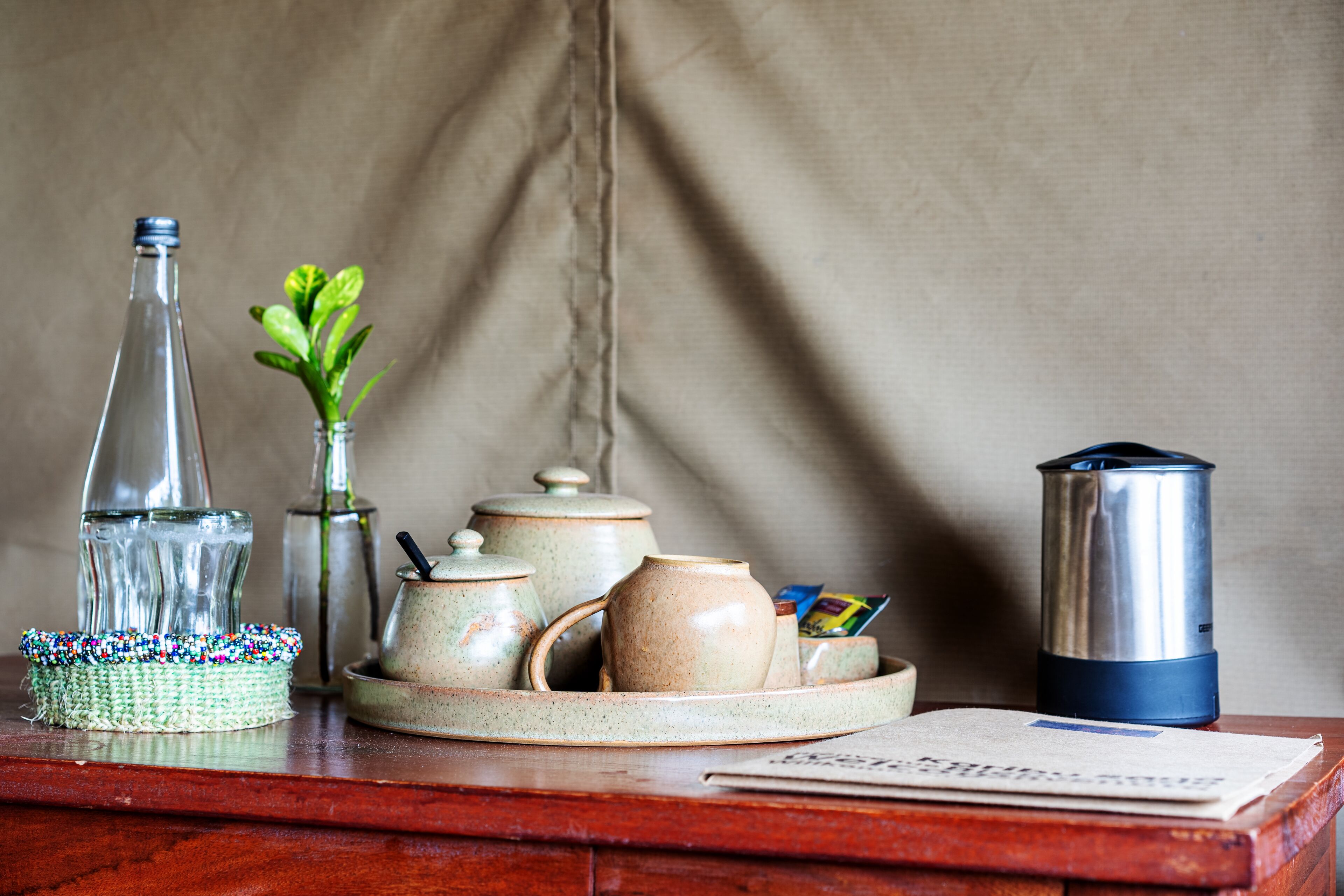 Una mesa de madera con un juego de té, una botella de vidrio, una planta pequeña y una cafetera.