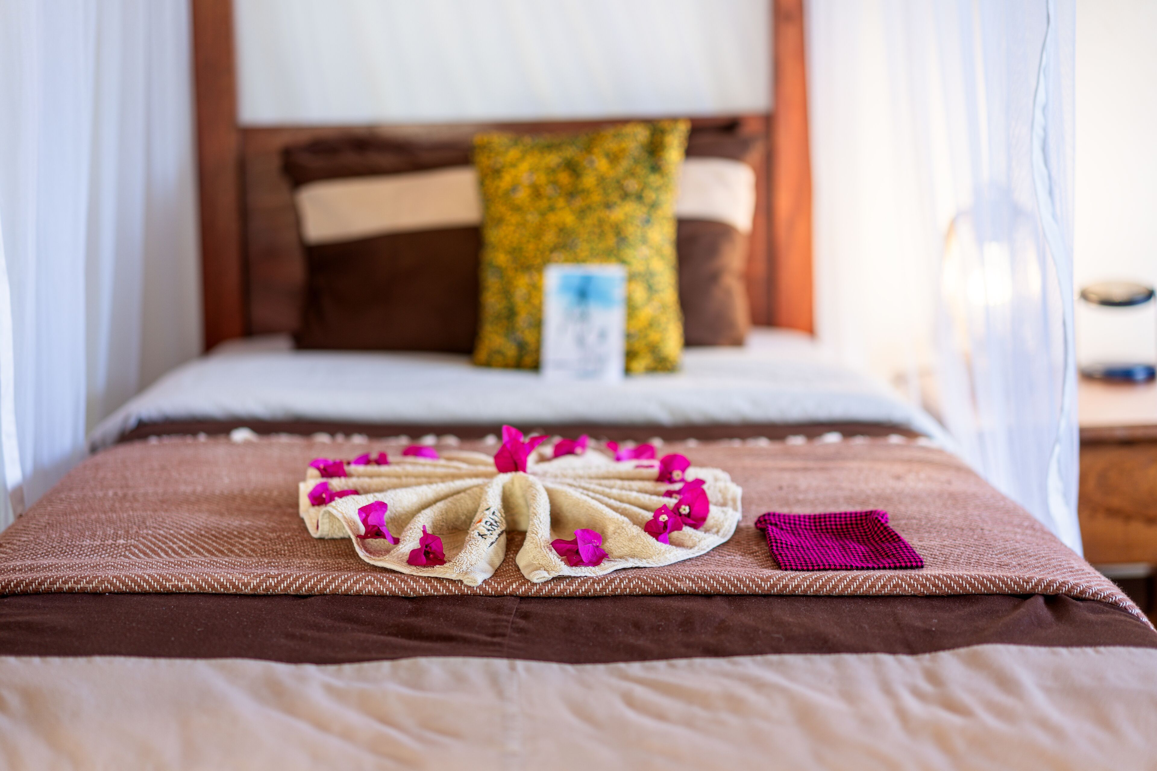 Uma cama bem arrumada com colcha marrom, arranjo de toalhas decorativo com flores rosa e almofada estampada em amarelo.
