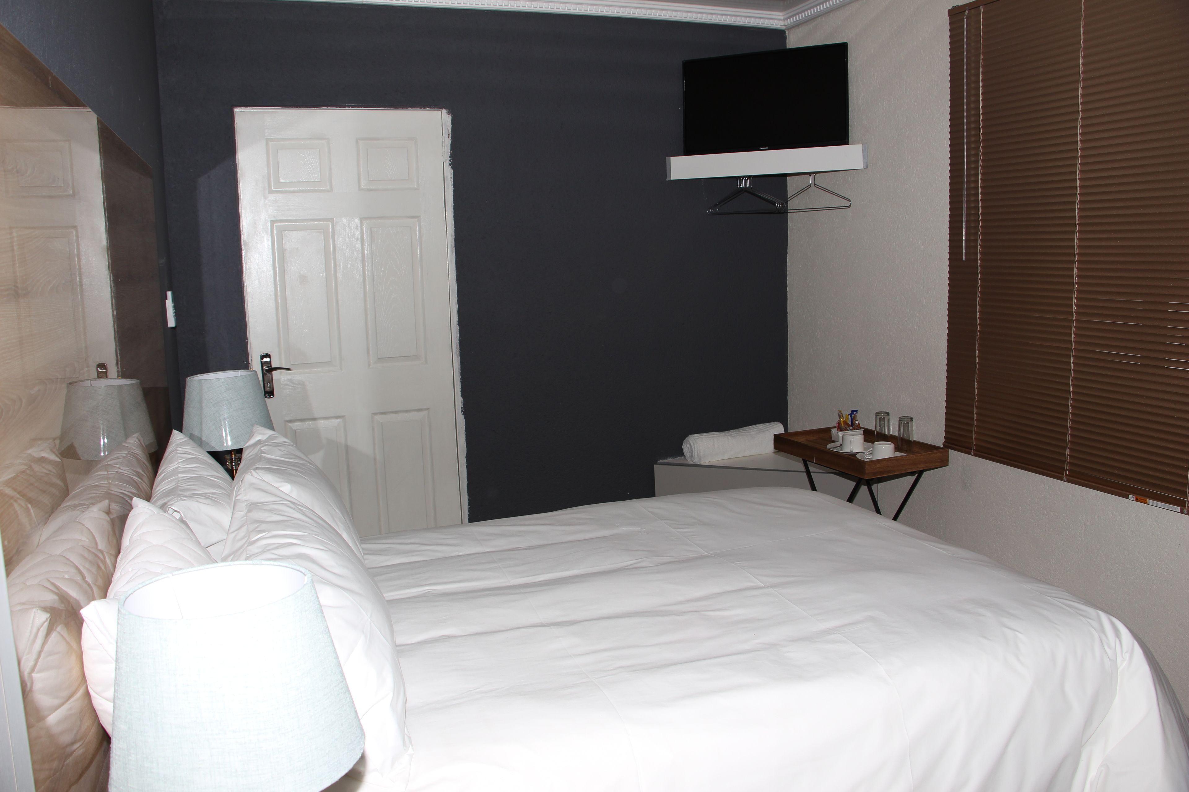 A hotel room with a bed, a bedside table, a TV mounted on the wall, and a window with blinds.