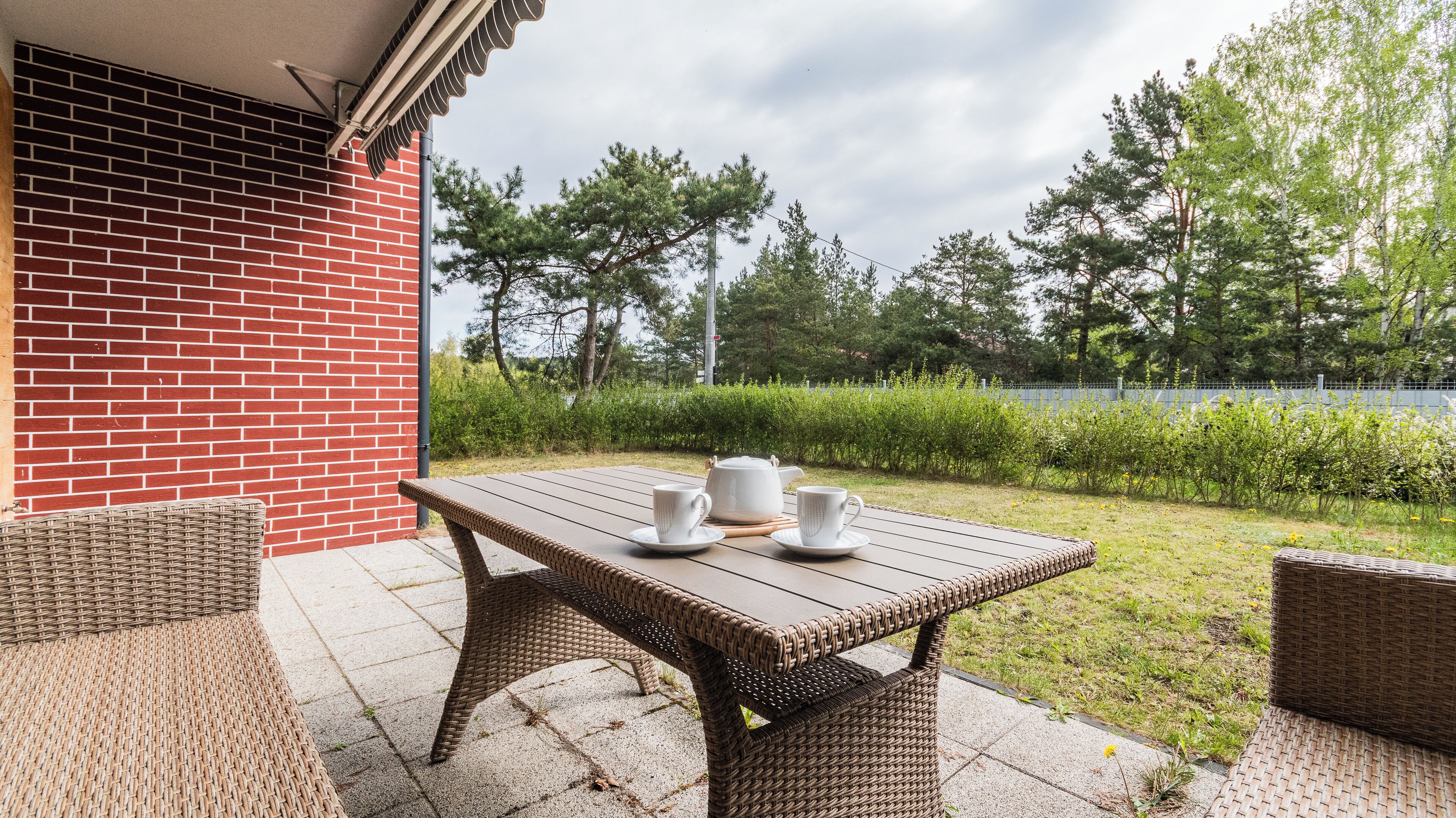 An outdoor patio with a wicker table set for two, two white cups, and a teapot.