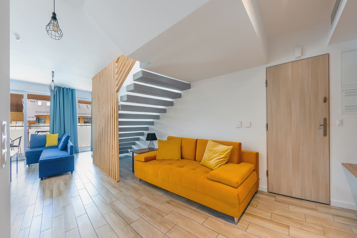 A modern living room with a staircase, a blue sofa, a yellow sofa, and a wooden door.