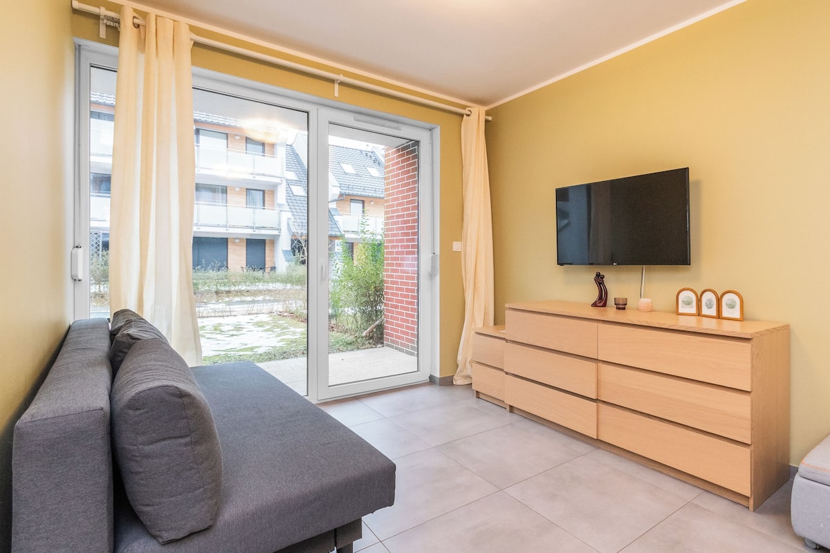 A modern living room with a sofa, a TV mounted on the wall, a wooden cabinet, and a sliding glass door leading to an outdoor area.