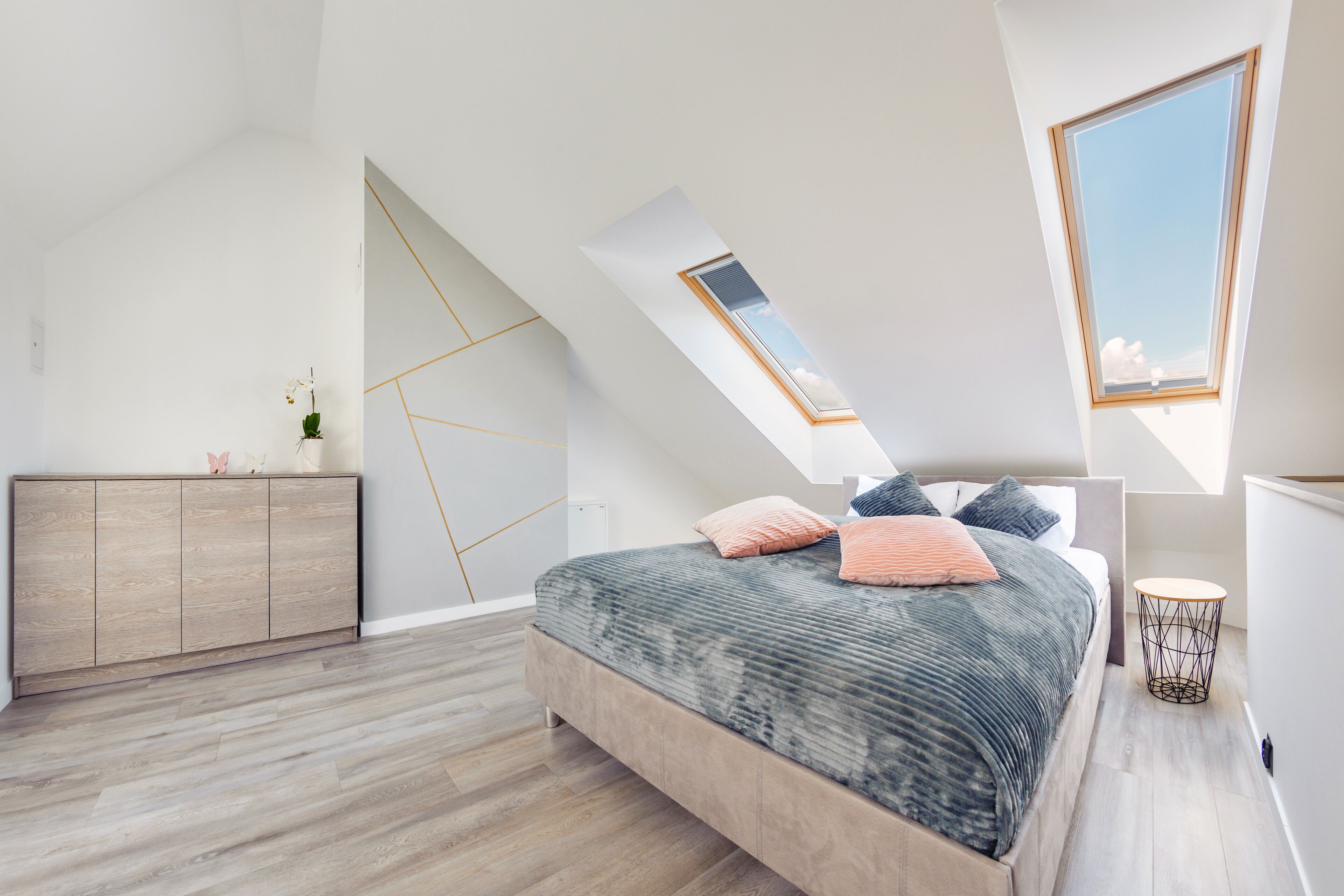 A modern bedroom with a sloped ceiling, a skylight, a bed with pillows, a bedside table, and a wooden cabinet.