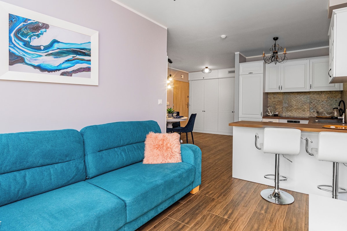A modern apartment with a teal sofa, a kitchen with white cabinets, and a dining area with a table and chairs.