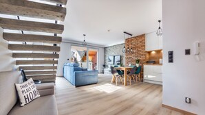 Un salón moderno con sofá, mesa de comedor y zona de cocina, con pared de ladrillo y suelo de madera.