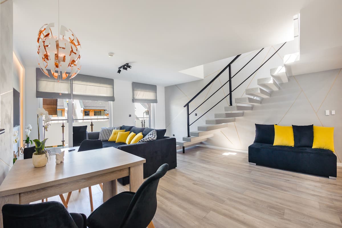 A modern living room with a wooden floor, a dining area with a table and chairs, a staircase, and a sofa with yellow cushions.
