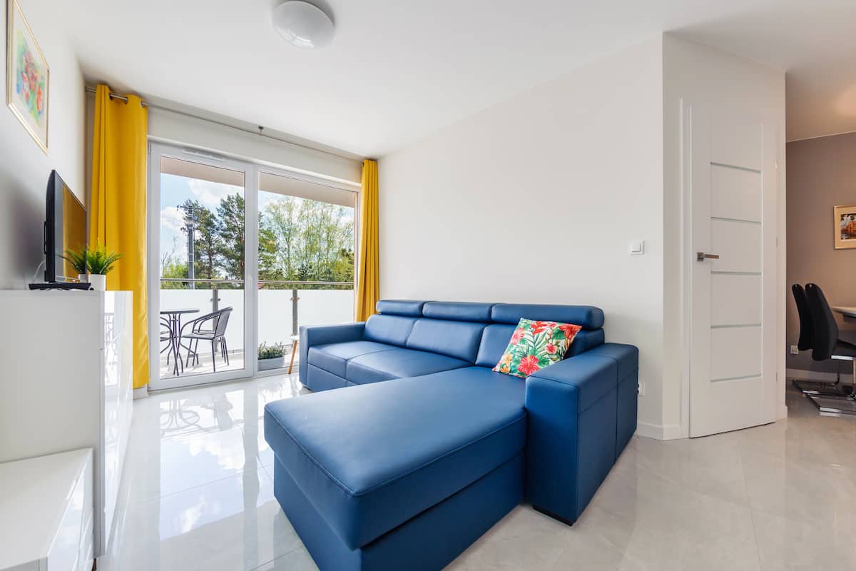 A modern living room with a blue sectional sofa, a flat-screen TV, and a balcony with yellow curtains.