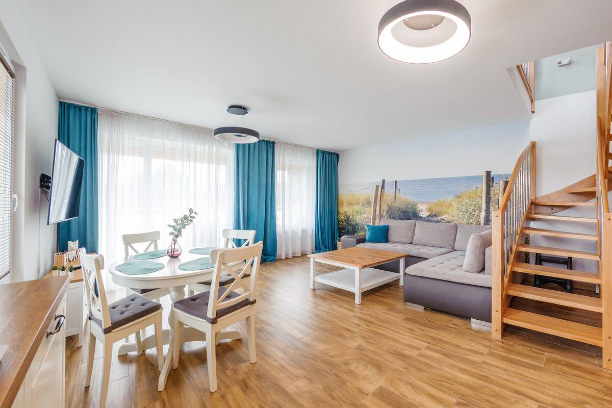A modern living room with a wooden staircase, a dining area with a table and chairs, and a large window with a view of the sea.