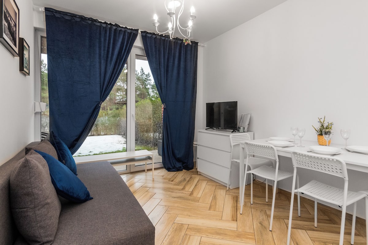 A modern living room with a sofa, a dining table with chairs, a TV, and a window with curtains.