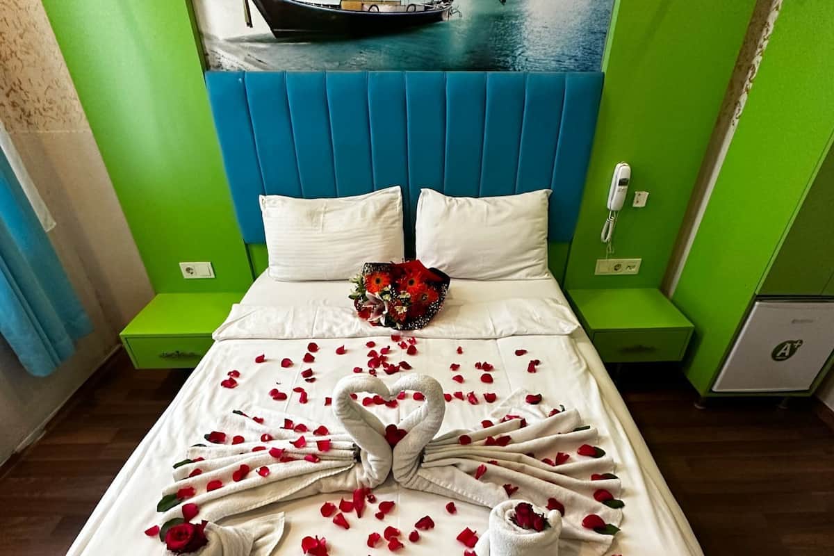 Un lit décoré de pétales de rose et de serviettes disposées en forme de cygne, un bouquet de fleurs rouges et un tableau représentant un bateau sur l’eau.