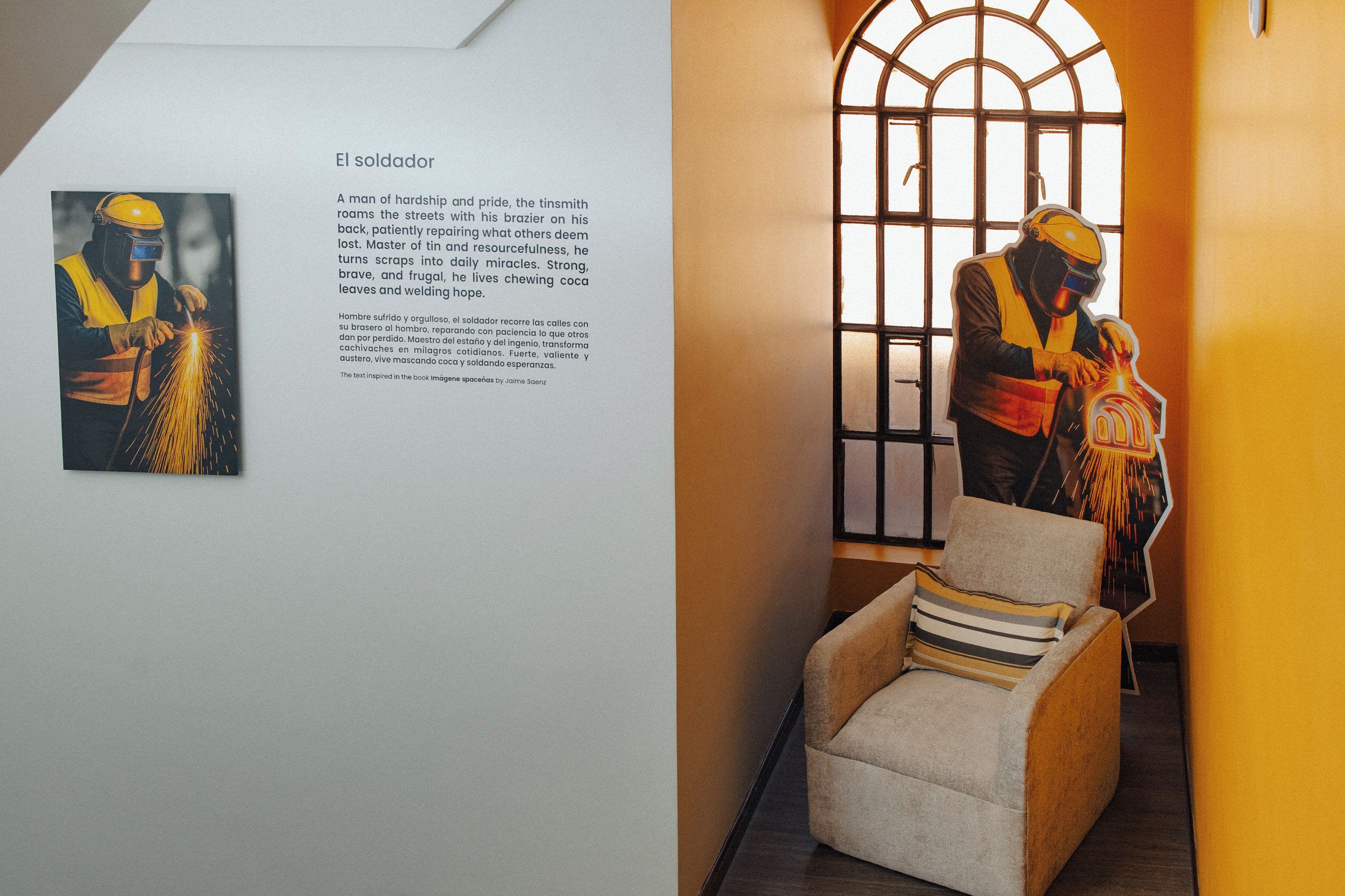 A corridor with a framed picture of a welder and text, a chair, and a window with a view.