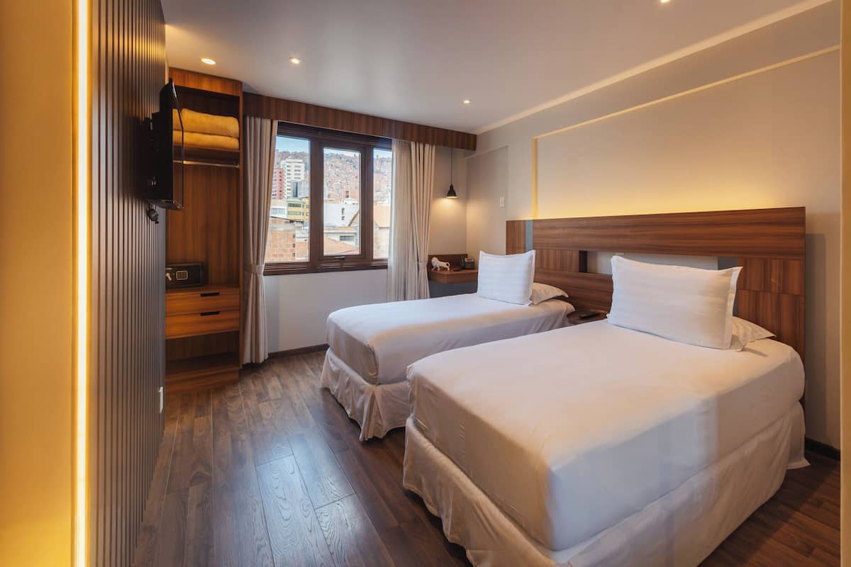Une chambre d’hôtel avec deux lits, un plancher en bois, une fenêtre avec des rideaux, une table de chevet et un téléviseur fixé au mur.