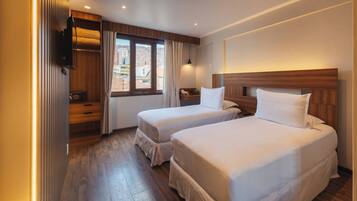 Habitación de hotel con dos camas, suelo de madera, una ventana con cortinas, una mesita de noche y un televisor de pared.