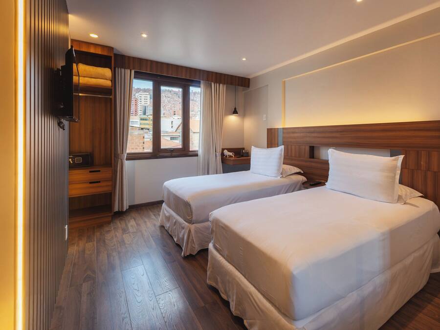 Habitación de hotel con dos camas, suelo de madera, una ventana con cortinas, una mesita de noche y un televisor de pared.