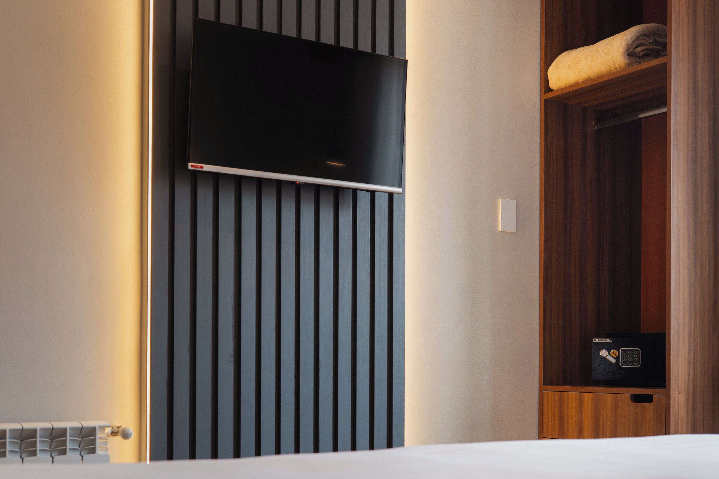 Una habitación de hotel moderna con un televisor de pantalla plana montado en una pared con listones verticales de madera, un armario de madera con caja fuerte y una cama bien hecha.