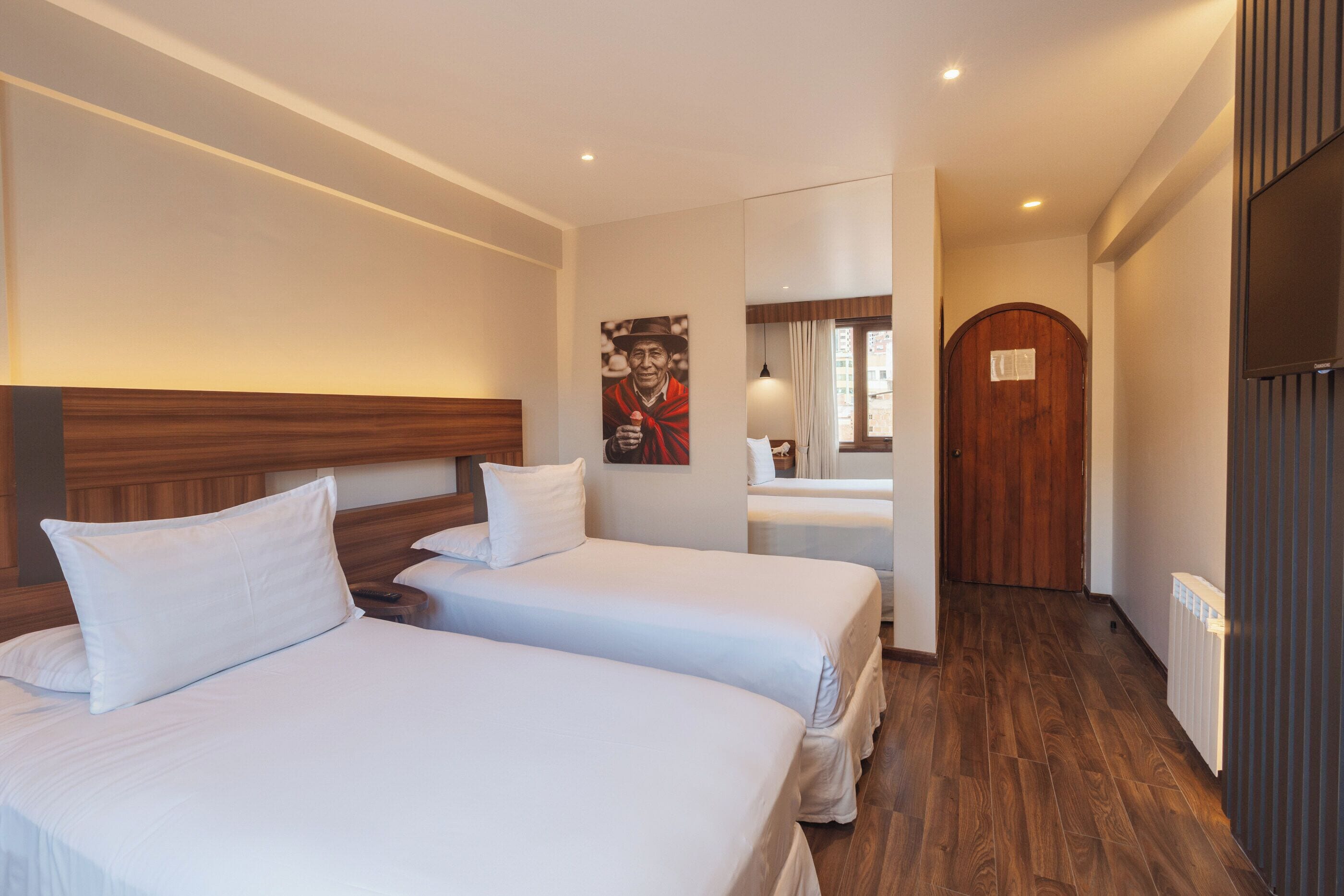 A hotel room with two beds, a wooden headboard, a poster on the wall, and a door leading to another room.
