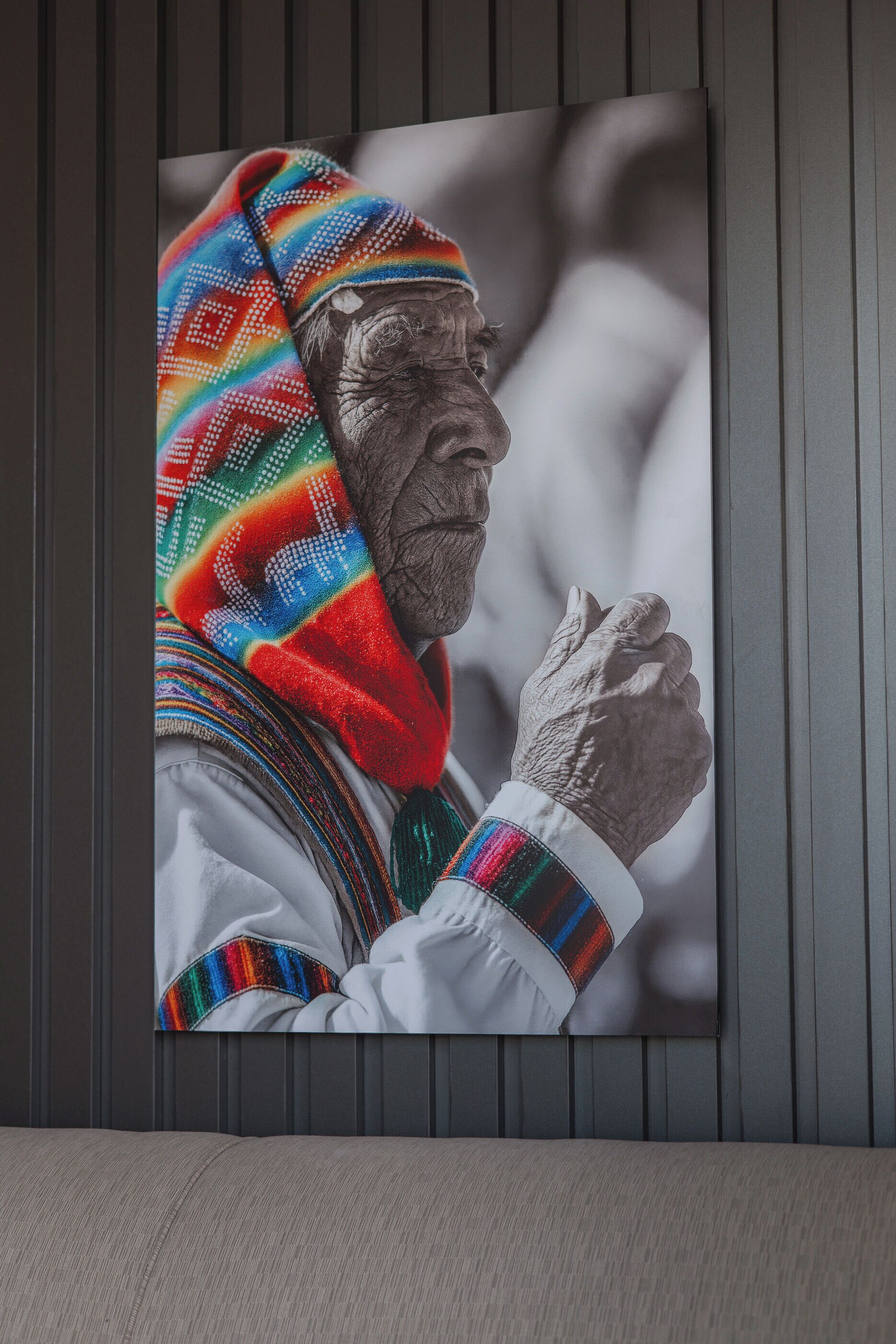 Een ingelijste foto van een persoon in traditionele kledij met een kleurrijk geborduurd sjaal, hangend aan een wand met verticale panelen.