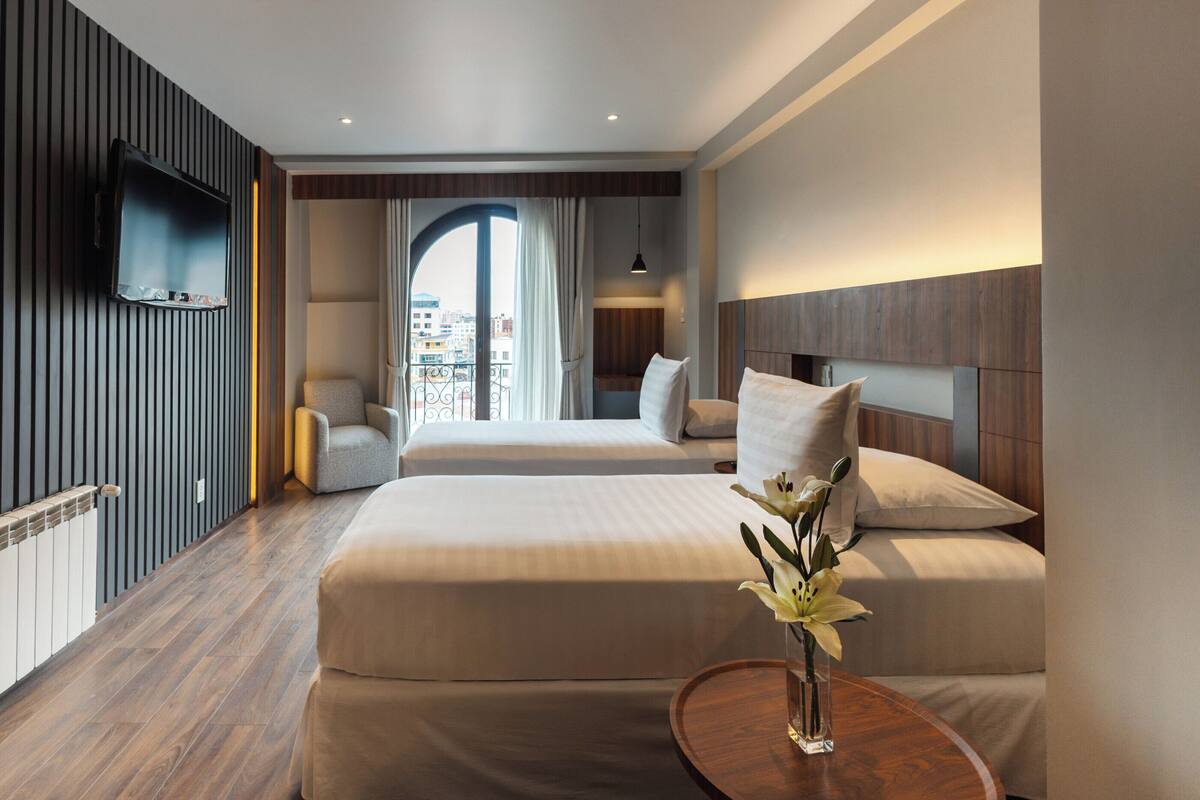 Une chambre d’hôtel moderne dotée d’un grand lit, d’un téléviseur fixé au mur, d’un petit coin salon et offrant une vue sur la ville grâce à une grande fenêtre.