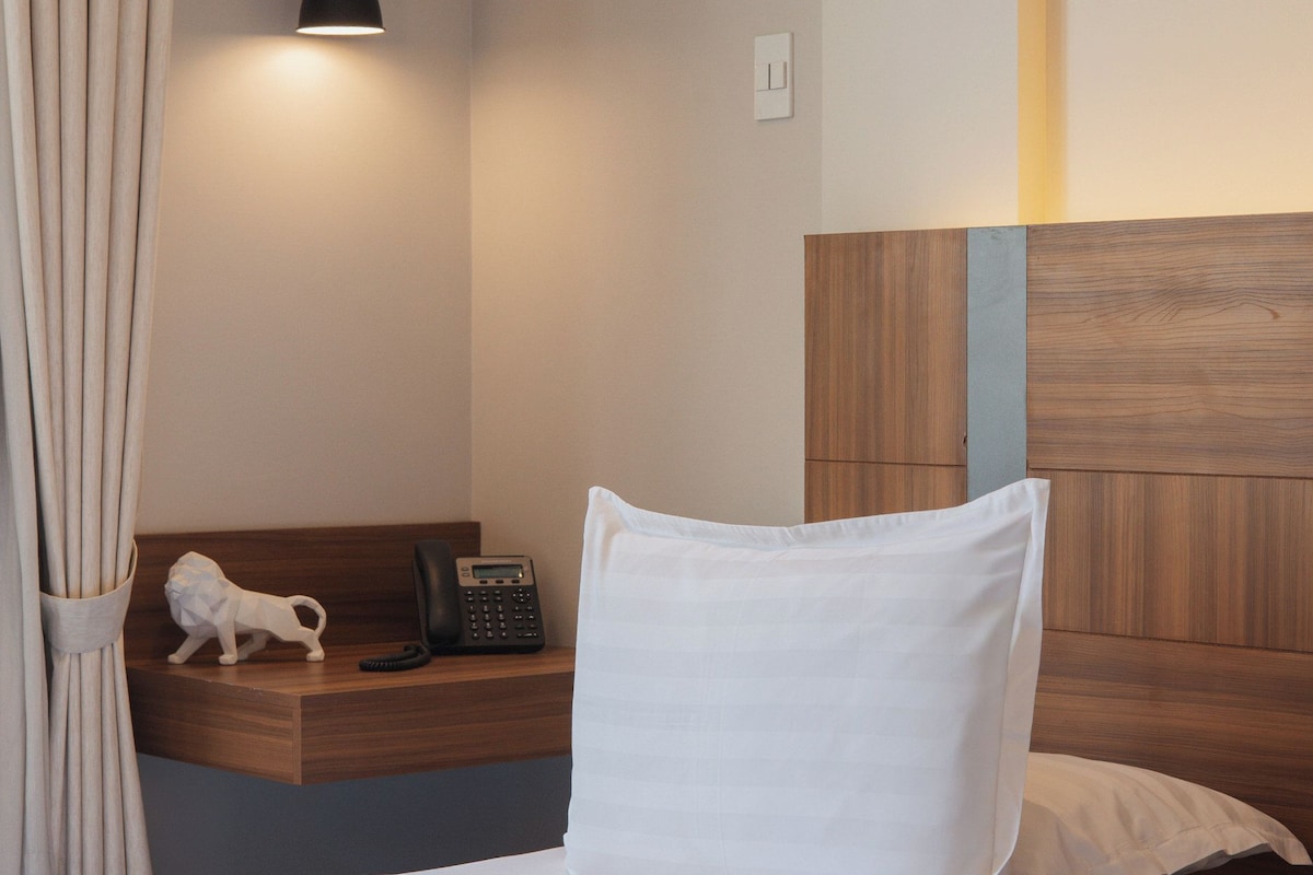 A hotel room with a bed, a bedside table with a phone, a lamp, and a curtain.