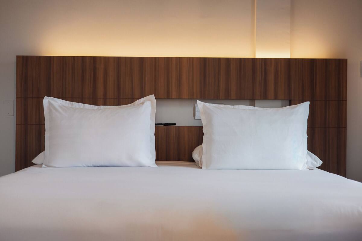 Un lit double avec du linge de lit blanc et deux oreillers, adossé à une tête de lit en bois dotée d’une étagère intégrée.