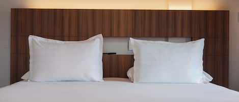 Una cama doble con sábanas blancas y dos almohadas, frente a un cabecero de madera con estante incorporado.