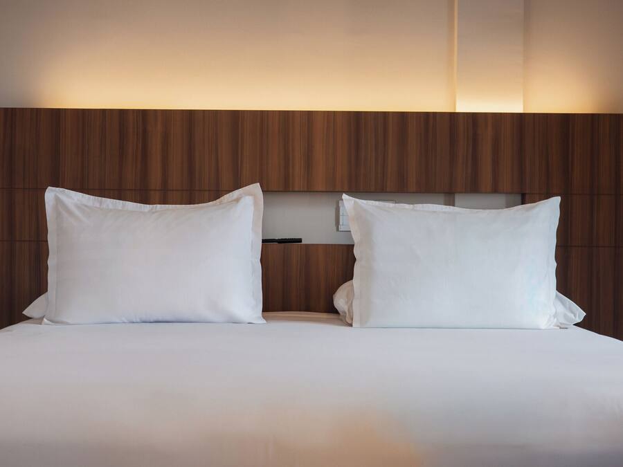 Una cama doble con sábanas blancas y dos almohadas, frente a un cabecero de madera con estante incorporado.