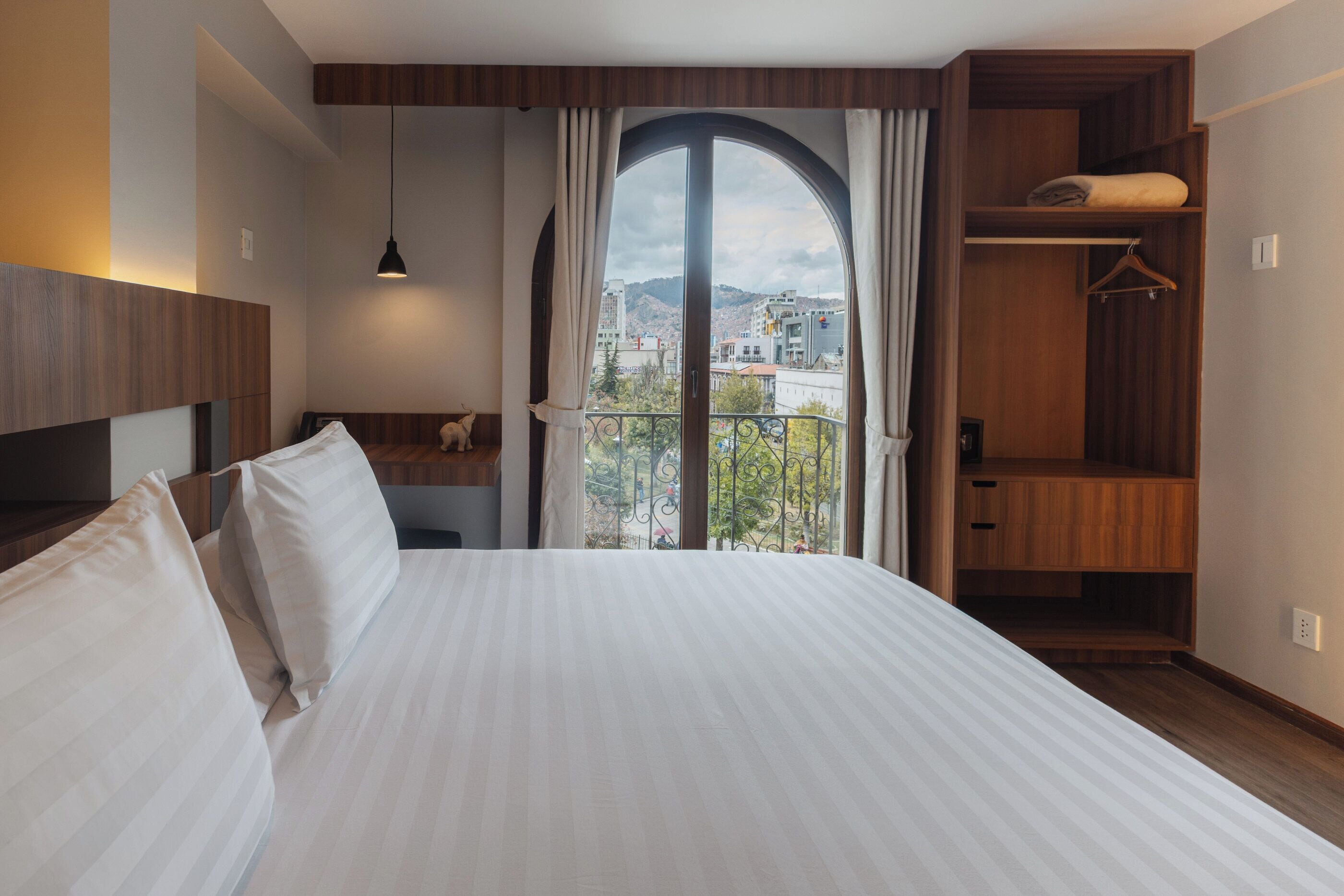 A hotel room with a large bed, a wooden wardrobe, a balcony with a view of buildings, and a hanging light fixture.